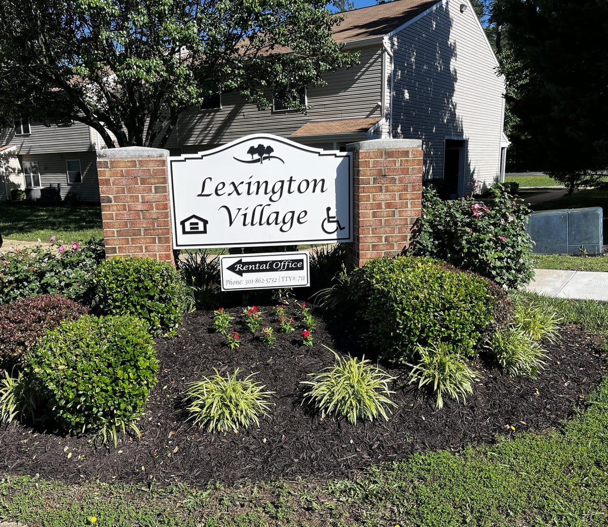 The Lexington Village entrance sign on brick pillars, framed by flower beds and shrubs, with a building behind on a sunny day