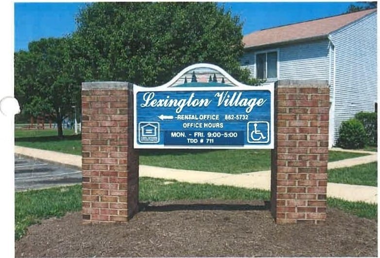 The Lexington Village entrance sign on brick pillars, with a building and trees behind on a sunny day